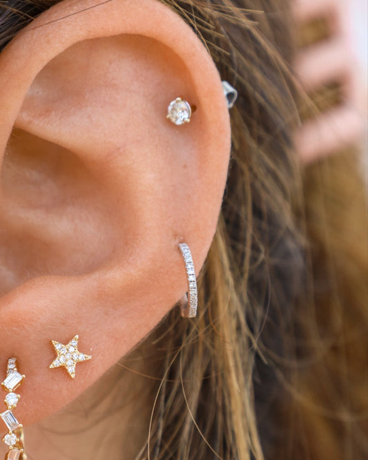 A close-up of a woman's ear wearing a small gold diamond hoop earring with multiple diamond accents.