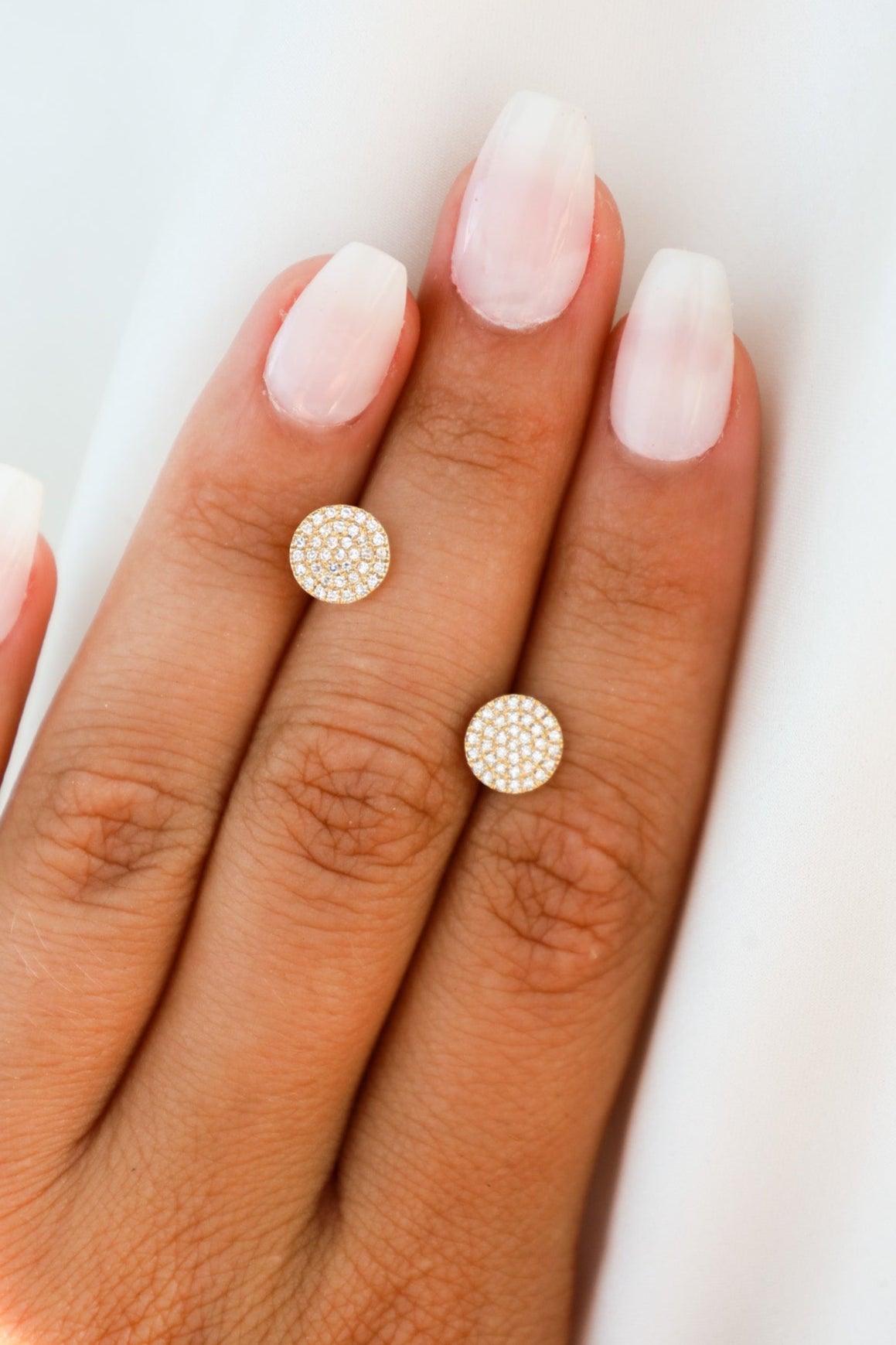 Gold stud earrings on a hand with light pink nail polish against a white background