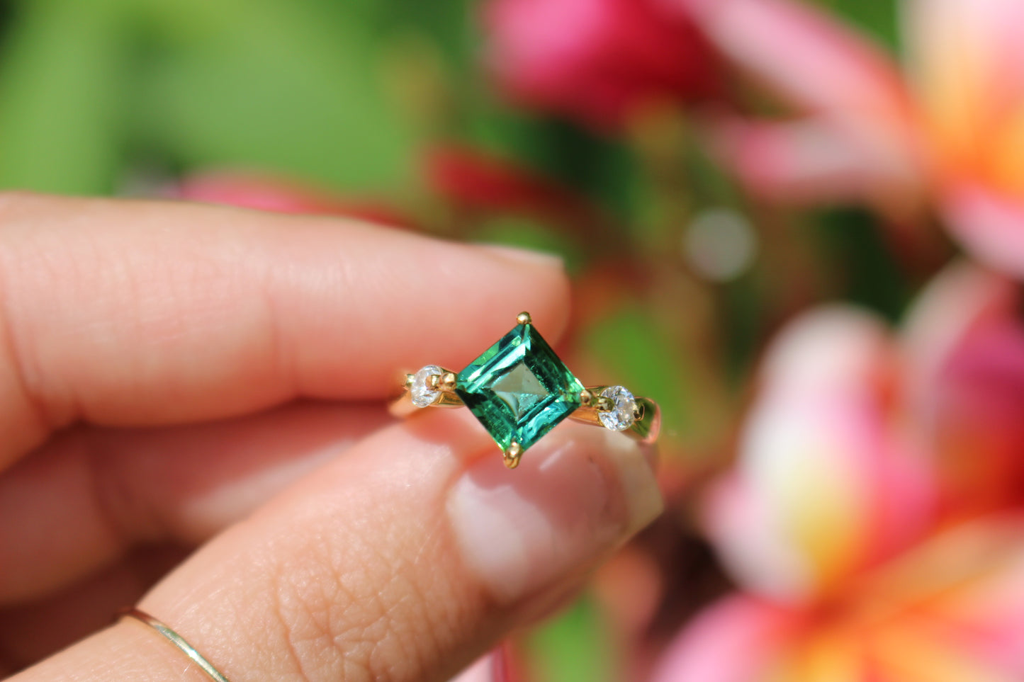 Green gemstone ring held by a hand with a blurred floral background