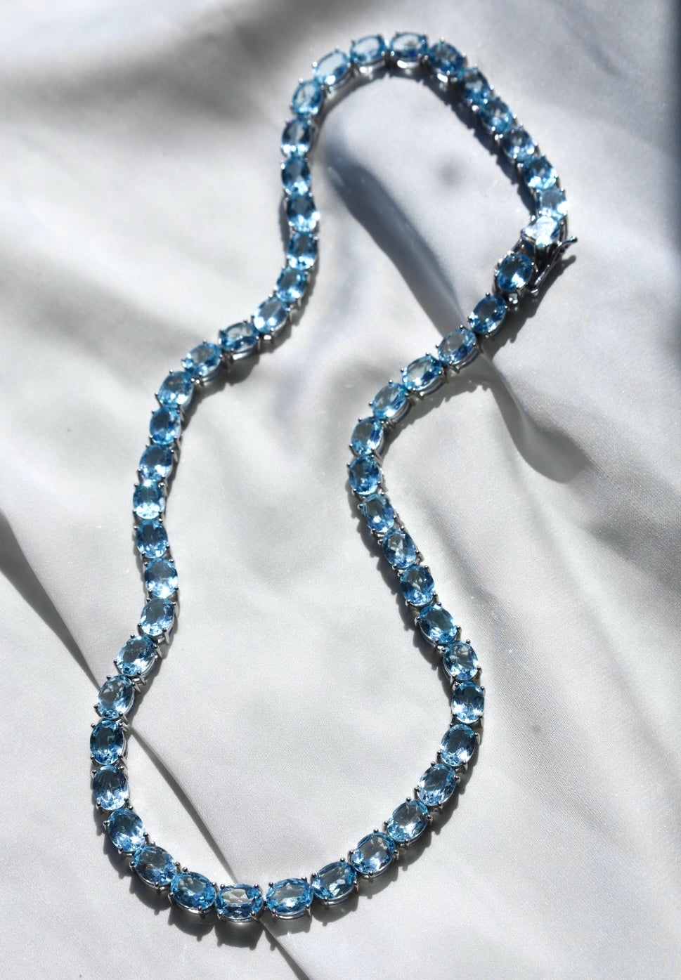A blue topaz tennis necklace with a silver clasp, displayed on a flat surface.