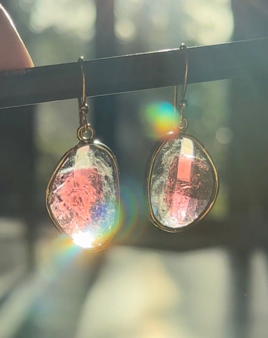Earrings with colorful, iridescent stones hanging on a black rod with a blurred natural background.