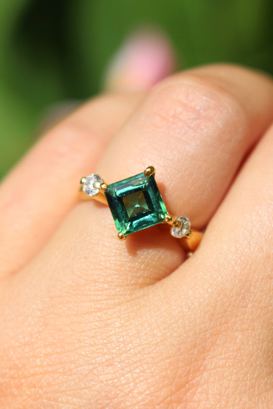 Gold ring with a green tourmaline on a person's finger against a blurred green background