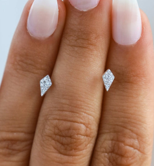 A person's hand showcasing a pair of diamond-shaped diamond stud earrings on a white background.