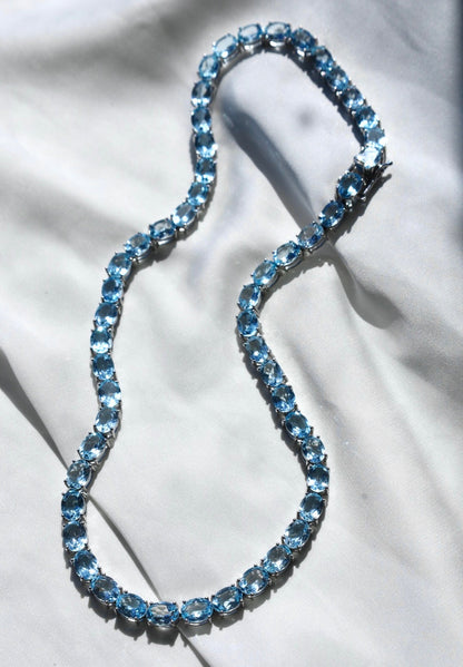 A blue topaz tennis necklace with a silver clasp, displayed on a flat surface.