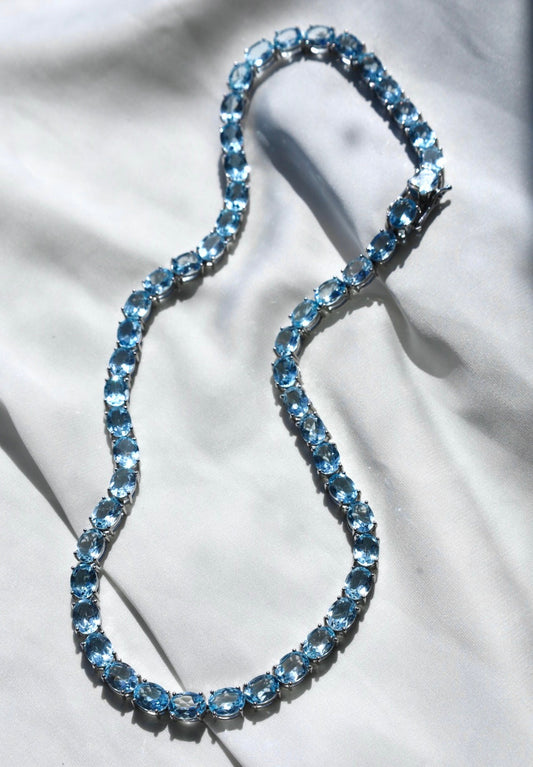A blue topaz tennis necklace with a silver clasp, displayed on a flat surface.