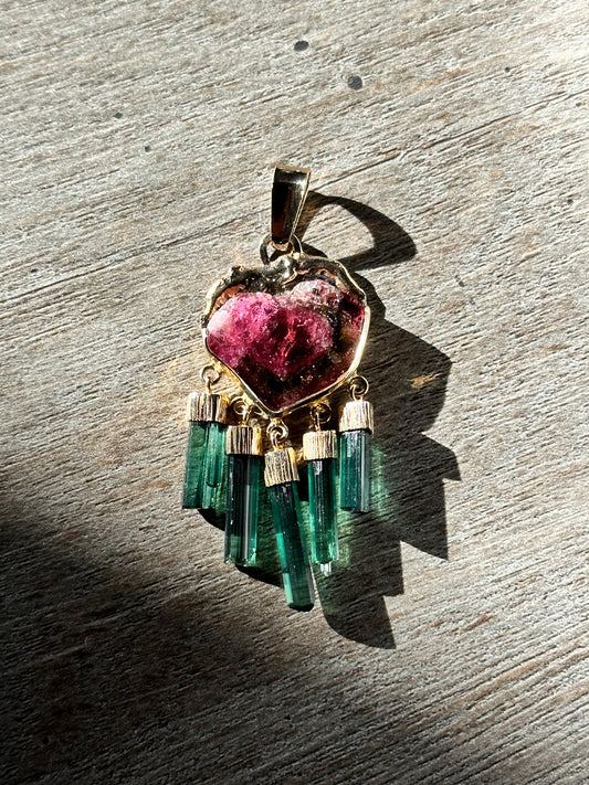 A 14K gold watermelon tourmaline pendant with indicolite crystals, featuring a polished surface and a heart-shaped design with a visible pink, green, and white color pattern.